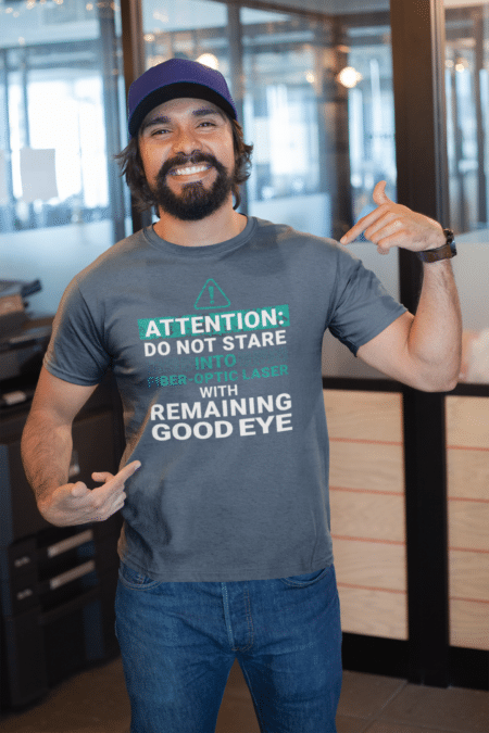 Man smiling and pointing to a t-shirt that says "Attention: do not stare into fiber-optic laser with remaining good eye"